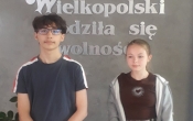 Nagrodzeni uczniowie Kaja i Patryk na tle szkolnego kacika patrona.